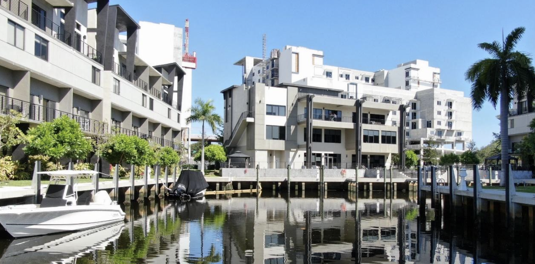 Photo of a modern waterfront building with a multi-story design, featuring balconies and large windows, located by a dock with palm trees in the foreground.