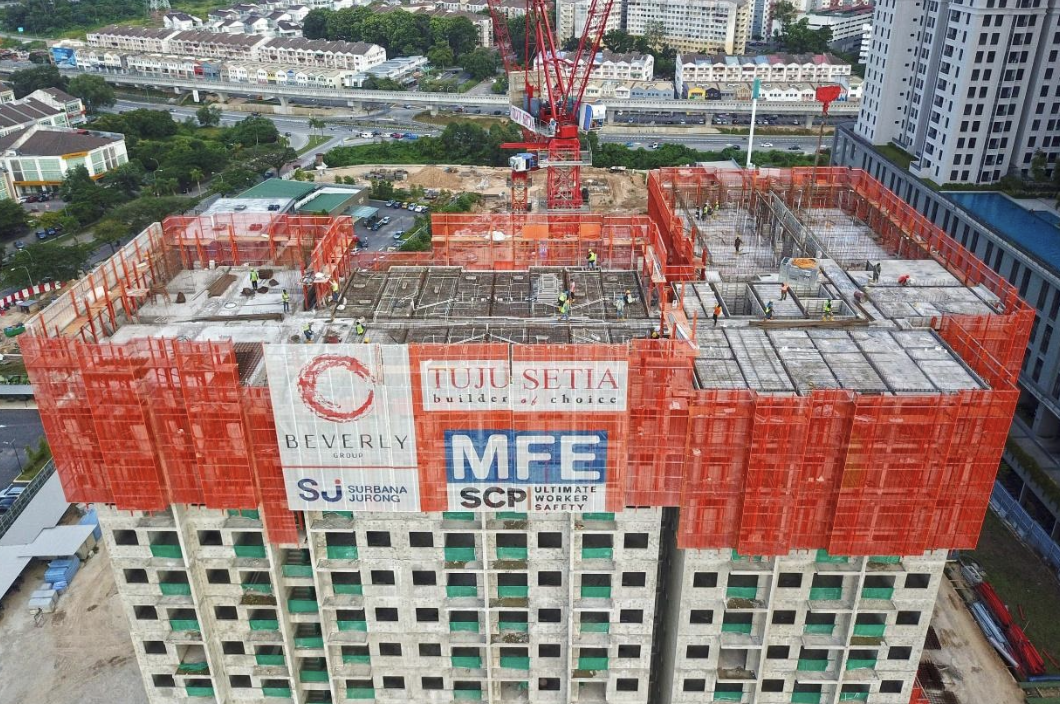 High-rise construction site with workers on the top floor, surrounded by orange safety netting and banners displaying contractor and project information.