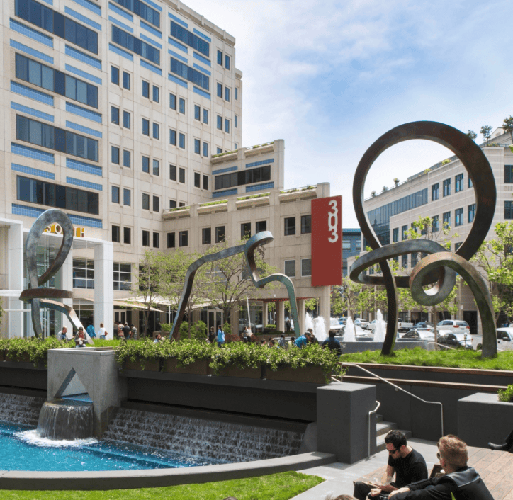 Urban plaza with abstract metal sculptures, fountain, and office buildings in the background.